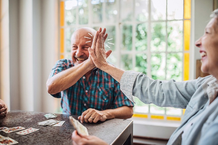 Older man high fiving a friend. They're sitting at a table playing cards.